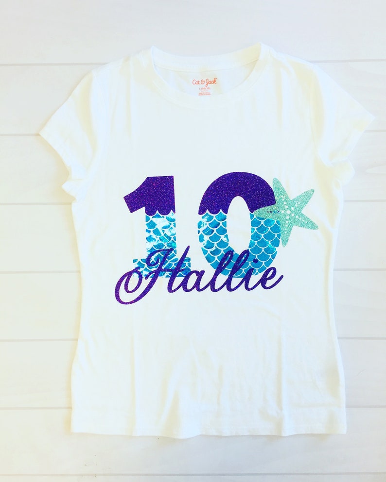 May include: White t-shirt with a purple and blue glitter number 10 with mermaid scales and a starfish. The name "Hallie" is written in purple glitter below the number.