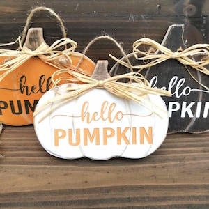 Könnte beinhalten: Drei hölzerne Kürbis-Ornamente mit dem Text "hello pumpkin" auf jedem. Die Kürbisse sind in Orange, Weiß und Schwarz bemalt mit einem braunen Stiel und einer Raffia-Schleife oben.