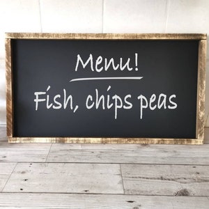 Pode incluir: Uma lousa preta com moldura de madeira, o texto "Menu!" escrito com giz branco, e o texto "Fish, chips peas" escrito com giz branco abaixo.
