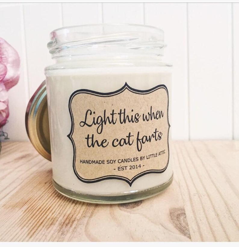 May include: A glass jar candle with a brown label that reads "Light this when the cat farts." The label also includes the text "Handmade soy candles by Little Attic - Est 2014 -."