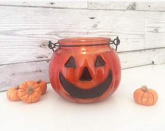 Könnte beinhalten: Orangener Glas-Kürbis-Kerzenhalter mit einem schwarzen Drahtgriff. Der Kerzenhalter hat ein geschnitztes Jack-o'-Lantern-Gesicht mit einem breiten Lächeln. Im Vordergrund befinden sich drei kleine orangefarbene Kürbisse.
