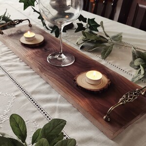Op de afbeelding: Een lange, rechthoekige houten serveerschaal met twee kleine kaarsen op houten rondjes en een wijnglas. De schaal heeft aan beide uiteinden een decoratief metalen handvat.