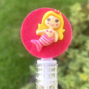 Op de afbeelding: Een bellenblaas met een roze vilten cirkel en een gele zeemeermin figuur. De zeemeermin heeft blond haar, een rode kroon en een roze en wit gestreepte staart. De transparante plastic buis is gevuld met bellen.