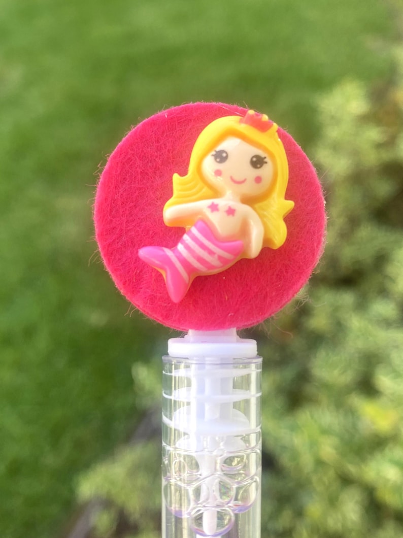 Op de afbeelding: Een bellenblaas met een roze vilten cirkel en een gele zeemeermin figuur. De zeemeermin heeft blond haar, een rode kroon en een roze en wit gestreepte staart. De transparante plastic buis is gevuld met bellen.