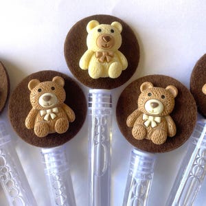May include: Bubble wands topped with brown felt circles and small teddy bear figurines. The bears are in shades of tan and cream, with bow ties. The wands are clear plastic.