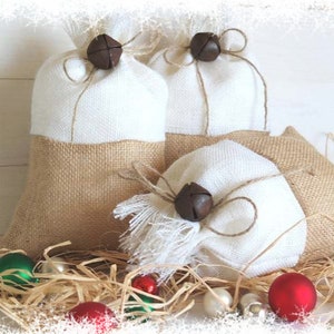 May include: Four white and burlap gift bags with brown bells tied with twine. The bags are sitting on a bed of straw with red and green Christmas ornaments.