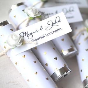 May include: White favor tubes decorated with gold hearts and topped with a white flower and a tag that reads "Megan & John Rehearsal Luncheon". The tubes have silver foil ends and are tied with twine.