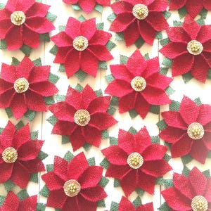 May include: A collection of handcrafted poinsettia flowers made from red and green burlap fabric. Each flower features layered petals and a central circular accent decorated with small, light-colored beads, arranged in a repeating pattern.
