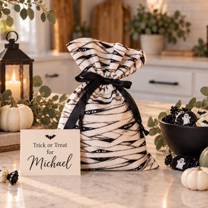 May include: A fabric drawstring bag featuring a mummy-inspired pattern of white bandages and black eyes, tied with a black ribbon. A rectangular card sits in front with the text Trick or Treat for Michael and a small black bat illustration.