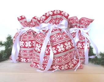 May include: Three red and white patterned fabric gift bags with white satin ribbon ties. The bags are arranged in a row on a white wooden surface.