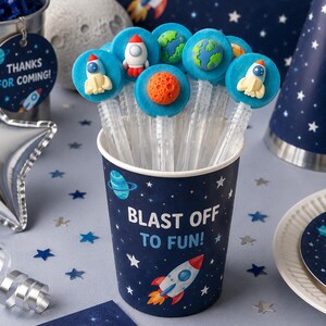 May include: A dark blue paper cup with the text BLAST OFF TO FUN! and rocket illustrations, filled with clear bubble wands. Each wand features a blue circular topper decorated with small 3D figures of rockets, planets, and moons.