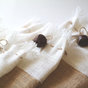 May include: Three white burlap gift bags with brown jingle bells tied with twine. The bags have a brown burlap bottom.