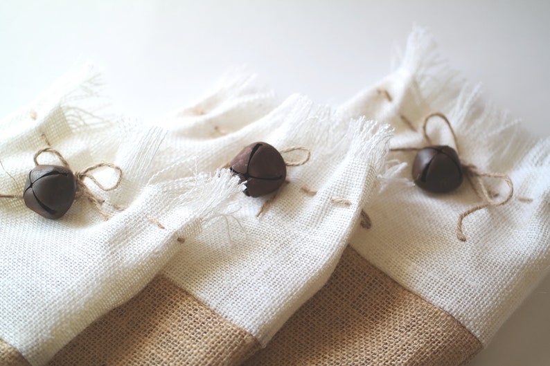 May include: Three white burlap gift bags with brown jingle bells tied with twine. The bags have a brown burlap bottom.