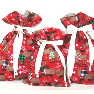May include: Three red fabric gift bags with white snowflake and moose designs. The bags are tied with white ribbon.
