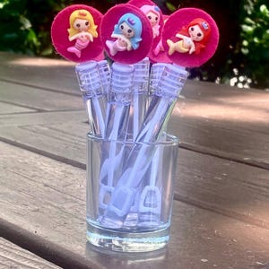 Op de afbeelding: Een verzameling bellenblaasstokken in een klein glas. Elke stok heeft een roze cirkel met een zeemeerminfiguur. De zeemeerminnen hebben verschillende haarkleuren: geel, blauw en rood. De stokken staan op een houten oppervlak.