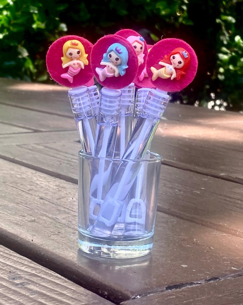 Op de afbeelding: Een verzameling bellenblaasstokken in een klein glas. Elke stok heeft een roze cirkel met een zeemeerminfiguur. De zeemeerminnen hebben verschillende haarkleuren: geel, blauw en rood. De stokken staan op een houten oppervlak.