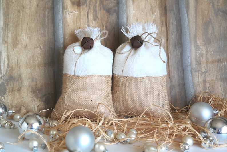 May include: Two small burlap gift bags with white fabric tops and brown bells tied with twine. The bags are sitting on a bed of straw with silver and white ornaments.