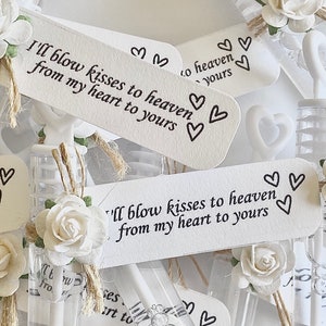 May include: White bubble wands with paper tags that read "I'll blow kisses to heaven from my heart to yours." Each wand is adorned with a white paper rose and twine. The wands are for a wedding or memorial.