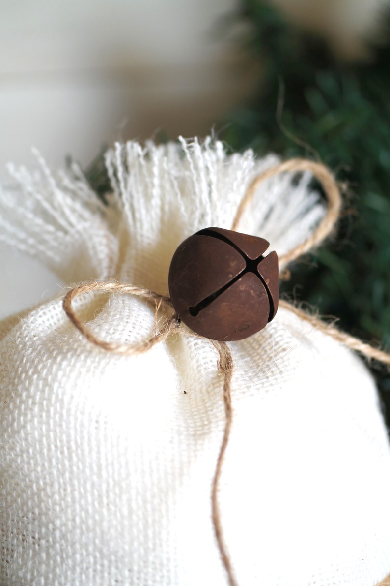 May include: A white burlap bag tied with twine and a rusty metal bell ornament. The bag is a rustic and simple gift wrap.
