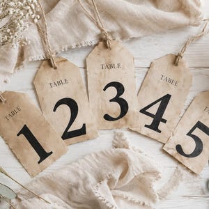 May include: Five rectangular paper tags with rounded top corners, each featuring the word TABLE and a large black number from one to five. The tags have a distressed, aged appearance with brown staining and are attached to natural jute twine strings.