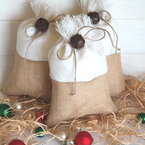 May include: Three burlap gift bags with white fabric tops, tied with twine and decorated with brown bells. The bags are sitting on a bed of shredded paper with red and green Christmas ornaments.