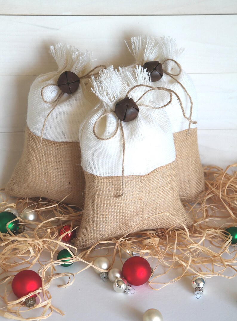 May include: Three burlap gift bags with white fabric tops, tied with twine and decorated with brown bells. The bags are sitting on a bed of shredded paper with red and green Christmas ornaments.