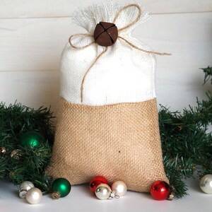 May include: A white and brown burlap gift bag with a brown bell tied with twine. The bag is surrounded by green pine branches and small Christmas ornaments in red, green, and white.