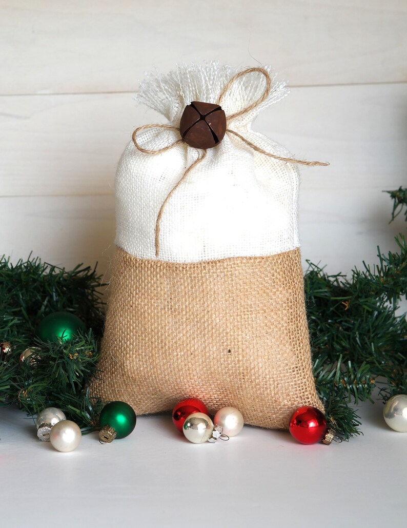 May include: A white and brown burlap gift bag with a brown bell tied with twine. The bag is surrounded by green pine branches and small Christmas ornaments in red, green, and white.