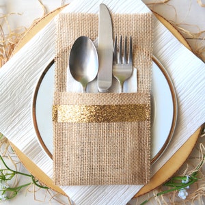 May include: A burlap napkin holder with a gold ribbon, holding a fork, knife, and spoon. The napkin holder is on a white textured placemat with a wooden base.
