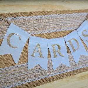 May include: A white banner with gold glitter letters spelling "CARDS" hangs on a burlap and lace background.