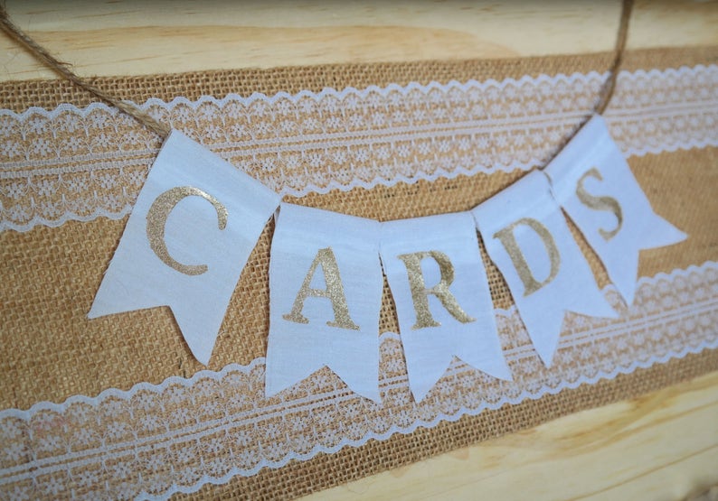 May include: A white banner with gold glitter letters spelling "CARDS" hangs on a burlap and lace background.