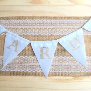 May include: White fabric bunting with gold letters spelling out the word "CARD" hanging on a burlap and lace background.