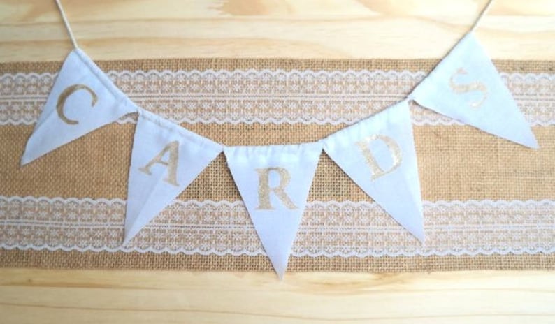 May include: White fabric bunting with gold letters spelling out the word "CARD" hanging on a burlap and lace background.