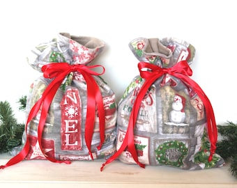May include: Two gray fabric gift bags with red ribbon bows. The bags have a Christmas theme with various holiday words and images printed on them. The bags are sitting on a wooden surface.