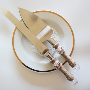 May include: A set of two silver cake knives with burlap and lace handles. The knives are on a white plate with a gold rim.