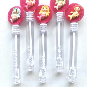 Op de afbeelding: Zes doorzichtige plastic bellenblaasstokken, bekroond met roze vilten cirkels en kleurrijke zeemeerminfiguren. Elke zeemeermin heeft een andere haarkleur: blauw, roze, geel, groen en rood. De stokken zijn verticaal gerangschikt.