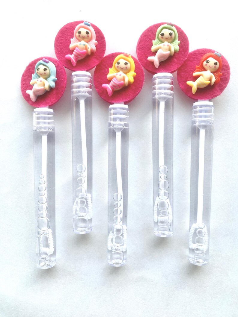 Op de afbeelding: Zes doorzichtige plastic bellenblaasstokken, bekroond met roze vilten cirkels en kleurrijke zeemeerminfiguren. Elke zeemeermin heeft een andere haarkleur: blauw, roze, geel, groen en rood. De stokken zijn verticaal gerangschikt.