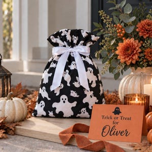 May include: A black fabric drawstring bag featuring a white ghost pattern, tied with a white satin ribbon. Next to it sits an orange rectangular card with a black ghost icon and the printed text: Trick or Treat for Oliver.