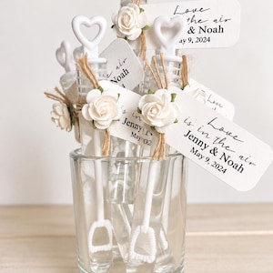May include: A collection of clear plastic bubble wands with white heart-shaped handles, each adorned with a small white fabric rose and a jute twine tie. Attached rectangular white tags feature the printed text Love is in the air, Jenny & Noah, May 9, 2024.
