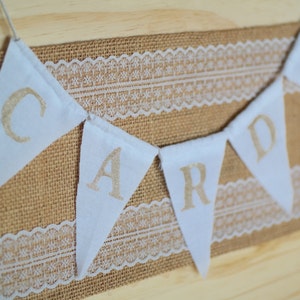 May include: A white banner with the word "CARDS" spelled out in gold letters. The banner is strung on a burlap background with white lace trim.