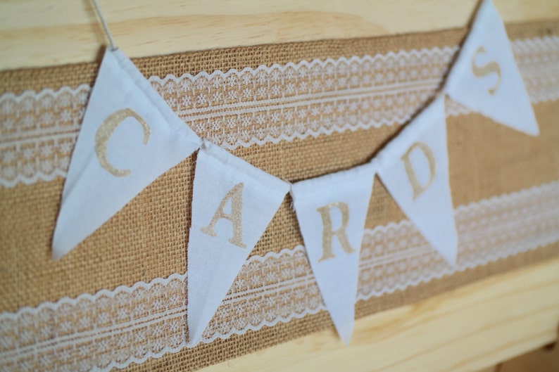 May include: A white banner with the word "CARDS" spelled out in gold letters. The banner is strung on a burlap background with white lace trim.