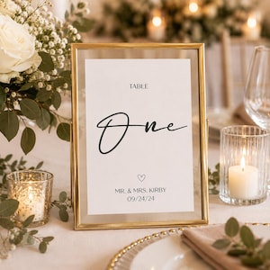 May include: A rectangular gold metal frame holding a white table number card. The card features the text TABLE One, a small heart icon, the names MR. & MRS. KIRBY, and the date 09/24/24 in black lettering against a clean white background.