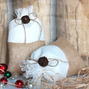 May include: Two white and burlap gift bags with brown bells tied with twine. The bags are sitting on a white surface with brown wood in the background. The bags are surrounded by brown shredded paper.
