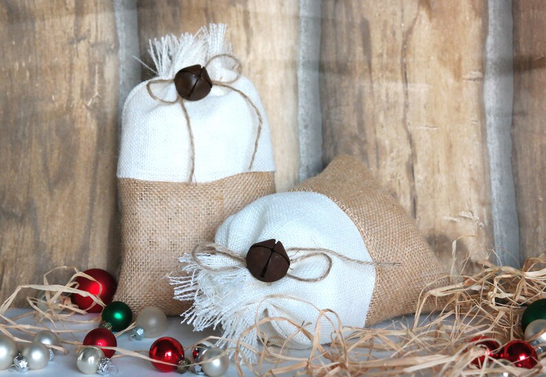 May include: Two white and burlap gift bags with brown bells tied with twine. The bags are sitting on a white surface with brown wood in the background. The bags are surrounded by brown shredded paper.