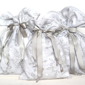 May include: Three white fabric gift bags with snowflake patterns and silver ribbon ties. The bags are sitting on a white surface.