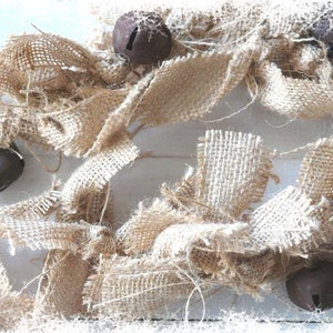May include: A rustic burlap garland with four dark brown metal bells. The garland is made of frayed burlap ribbon and is arranged in a decorative pattern. The bells are evenly spaced throughout the garland.