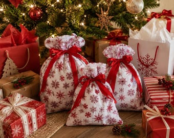 May include: Three white fabric gift bags featuring a red snowflake pattern, each cinched at the top with a vibrant red satin ribbon bow. These bags are surrounded by various wrapped presents, including red and white boxes with ribbons.