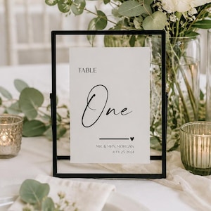 May include: A rectangular black metal frame holding a white card with black text that reads TABLE One, MR. & MRS. MORGAN, and JULY 25, 2024. The card features a small black heart icon and a horizontal line separating the text elements.