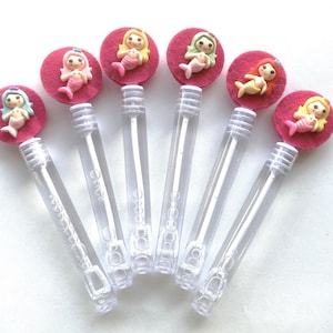 Op de afbeelding: Bellenstokken met zeemeermin-toppers. Elke toverstaf heeft een doorzichtige plastic buis met een bellenoplossing en een decoratieve bovenkant. De toppers zijn roze vilten cirkels met een zeemeerminfiguur in verschillende kleuren.