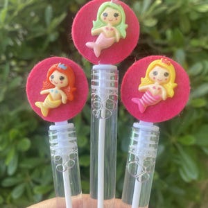 Op de afbeelding: Drie bellenblaasstokken met zeemeermin-toppers. Elke toverstaf heeft een doorzichtige plastic buis met een witte toverstaf en een roze vilten cirkel met een zeemeerminfiguur. De zeemeerminnen hebben verschillende haar- en staartkleuren: groen, oranje en geel.
